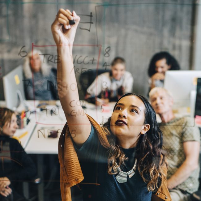 Businesswoman explaining strategy to colleagues while writing on glass at IT company